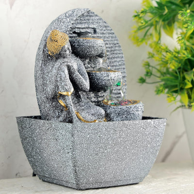A water fountain featuring a statue of Lord Buddha, the founder of Buddhism, surrounded by cascading waterfalls or a serene pool of water. The fountain may be made of stone, marble, or other materials and may have additional decorative elements, such as plants or lights.