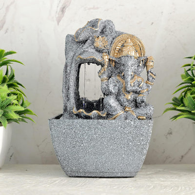 A water fountain featuring a statue of Lord Ganesha, the Hindu god of wisdom and remover of obstacles. Water flows around or from the statue, creating a serene and calming atmosphere. The fountain may be made of stone, marble, or other materials and may have additional decorative elements, such as plants or lights