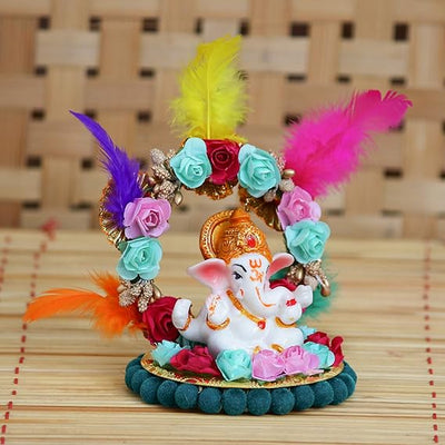 A small golden idol of Lord Ganesha, the Hindu god of wisdom and remover of obstacles, seated on a throne made of colorful flowers. The idol is placed on a car dashboard.