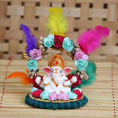 A small golden idol of Lord Ganesha, the Hindu god of wisdom and remover of obstacles, seated on a throne made of colorful flowers. The idol is placed on a car dashboard.