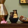 A water fountain featuring a statue of Buddha seated in meditation pose. Water flows from the Buddha's hands or from a nearby source, creating a serene and calming atmosphere. The fountain may be made of stone, marble, or other materials and may have additional decorative elements.