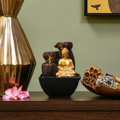 A water fountain featuring a statue of Buddha seated in meditation pose. Water flows from the Buddha's hands or from a nearby source, creating a serene and calming atmosphere. The fountain may be made of stone, marble, or other materials and may have additional decorative elements.