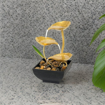 A small, tabletop fountain made of metal. Water flows through the metal pipes or channels, creating a soothing sound. The fountain may have additional decorative elements, such as rocks, plants, or a figurine.