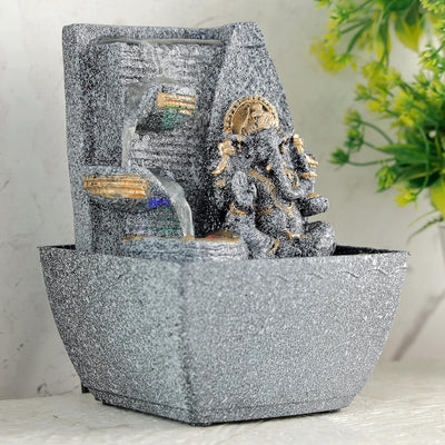 A small, tabletop fountain made of polyresin, a durable and lightweight material, featuring a statue of Lord Ganesha, the Hindu god of wisdom and remover of obstacles. Water flows around or from the statue, creating a serene and calming atmosphere. The fountain may have additional decorative elements, such as plants or lights.