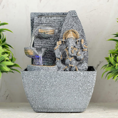 A small, tabletop fountain made of polyresin, a durable and lightweight material, featuring a statue of Lord Ganesha, the Hindu god of wisdom and remover of obstacles. Water flows around or from the statue, creating a serene and calming atmosphere. The fountain may have additional decorative elements, such as plants or lights.