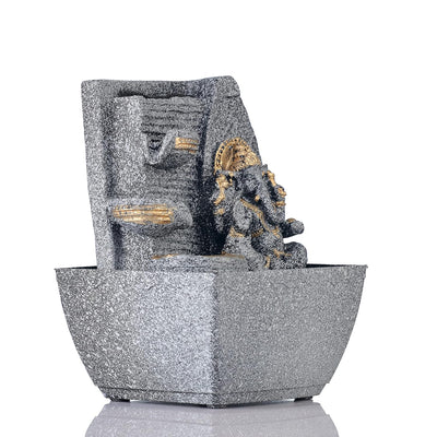 A small, tabletop fountain made of polyresin, a durable and lightweight material, featuring a statue of Lord Ganesha, the Hindu god of wisdom and remover of obstacles. Water flows around or from the statue, creating a serene and calming atmosphere. The fountain may have additional decorative elements, such as plants or lights.