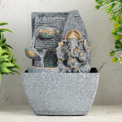 A small, tabletop fountain made of polyresin, a durable and lightweight material, featuring a statue of Lord Ganesha, the Hindu god of wisdom and remover of obstacles. Water flows around or from the statue, creating a serene and calming atmosphere. The fountain may have additional decorative elements, such as plants or lights.