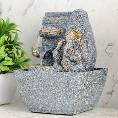 A small, tabletop fountain made of polyresin, a durable and lightweight material, featuring a statue of Lord Ganesha, the Hindu god of wisdom and remover of obstacles. Water flows around or from the statue, creating a serene and calming atmosphere. The fountain may have additional decorative elements, such as plants or lights.
