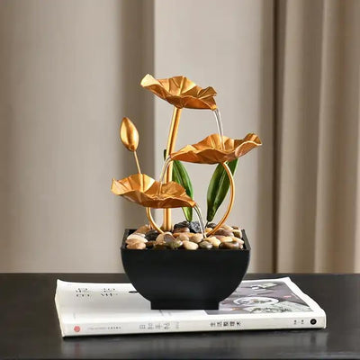 A small, tabletop fountain featuring a lotus flower design. Water flows from the lotus petals or from a nearby source, creating a serene and calming atmosphere. The fountain may be made of ceramic, stone, or other materials and may have additional decorative elements.