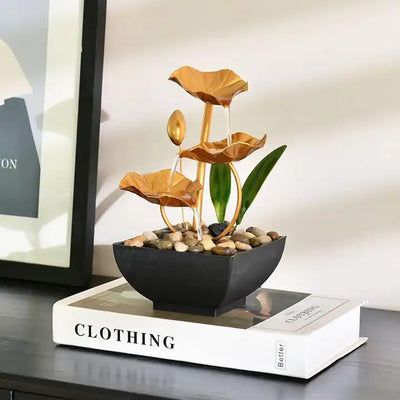 A small, tabletop fountain featuring a lotus flower design. Water flows from the lotus petals or from a nearby source, creating a serene and calming atmosphere. The fountain may be made of ceramic, stone, or other materials and may have additional decorative elements.